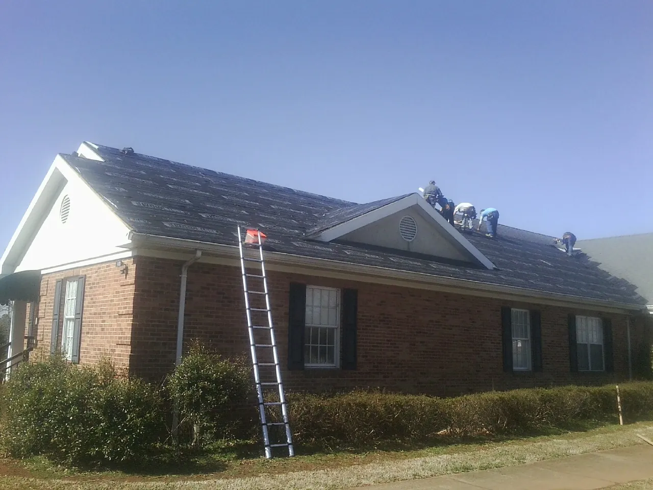 Crew tearing shingles off a brick ranch roof with a ladder set — Emergency Roof Repair work by our crew in Grenada