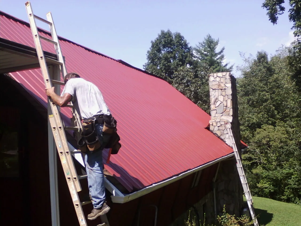 Roofer on a ladder beside a red metal roof and stone chimney — Soffit & Fascia Repair by Roofing Brothers in Grenada